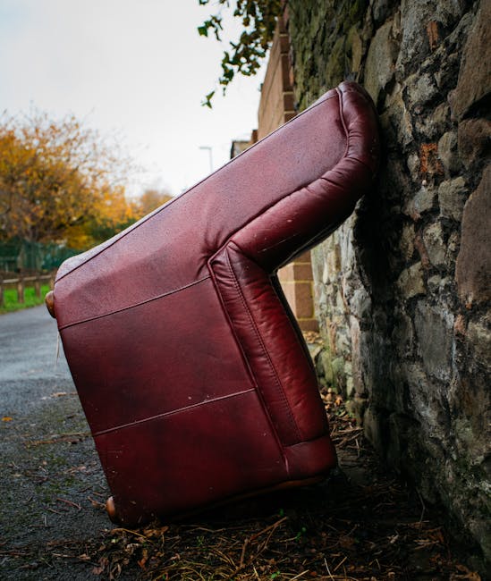 A large, burgundy leather armchair with visible stitching rests at an angle against a rough stone wall outside a residential property. The armchair is positioned on a narrow pavement near the edge of a road, with some fallen leaves and small debris around its base. The background features an overcast sky, a green fence, and autumn foliage on nearby trees, indicating a typical residential setting in HERNE HILL. The scene captures an ongoing home relocation or furniture transport process, with the armchair temporarily outside, possibly awaiting collection or reloading by Man with Van Herne Hill for a house removal service.