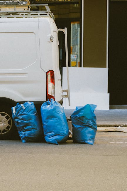 A white commercial van parked on a street outside a building with large glass windows, partially visible doors, and a white tiled step ledge. The van's rear door is open, revealing a glimpse of the vehicle's interior. Three large blue plastic bags filled with household items or packaging materials are placed on the pavement next to the van, positioned towards the left side of the image. The scene appears to be part of a home relocation or furniture transport process, with the bags likely containing items prepared for loading or unloading. The lighting suggests daylight, and the setting indicates an outdoor loading area for a professional removal service, with the company's name and service details available on the associated webpage.