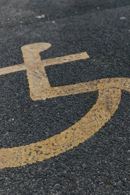 Close-up view of a yellow-painted disabled accessible parking symbol on an asphalt surface, with visible cracks and texture. The symbol's lines are slightly worn, indicating regular use. The image captures the parking area adjacent to a property, likely part of the access point for a home or commercial building involved in a nearby house removal or relocation process. No vehicles or other objects are visible in the frame. This type of marking is common in urban settings to designate spaces for relocating furniture or belongings during home relocation services provided by Man with Van Herne Hill. The lighting appears natural, suggesting daytime conditions, and the surface is clean without debris. The focus is on the parking designation, supporting the context of moving logistics and accessible parking during a professional removal service at Croxted Road.