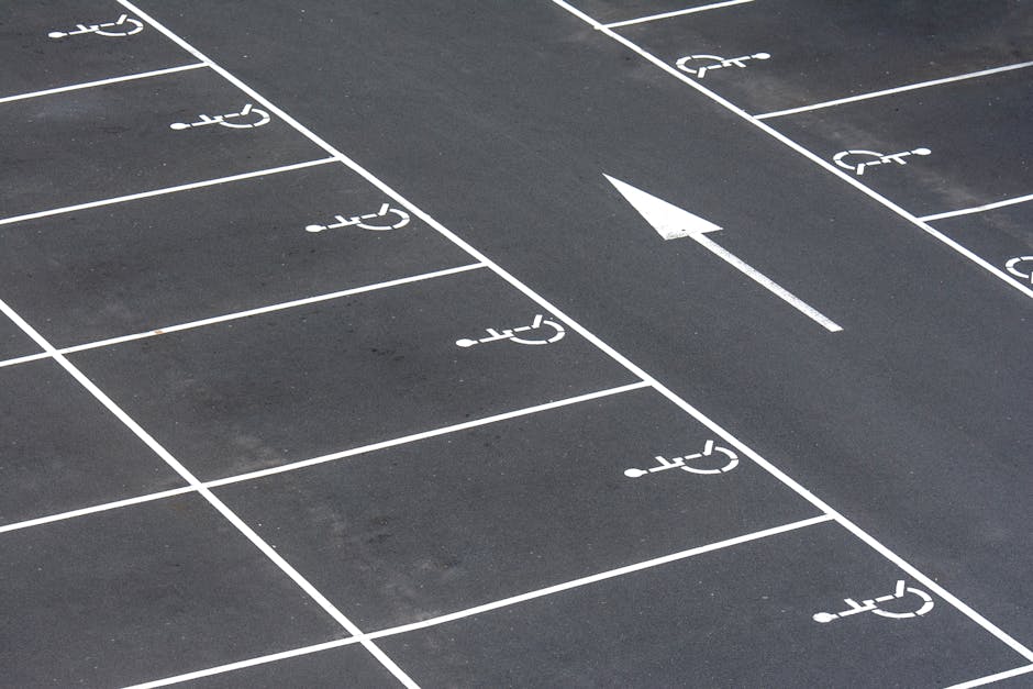 A section of an empty parking lot with designated spaces marked by white lines, including several wheelchair accessible parking bays with the wheelchair symbol painted in each space. The surface is made of dark asphalt, and a single white arrow painted on the ground indicates the direction of traffic flow within the lot. The image captures an unobstructed view focusing on the parking layout, highlighting accessible parking areas, and providing context for vehicle and equipment movement during house removals or home relocation activities, similar to those described on the Croxted Road Removals page by Man with Van Herne Hill.