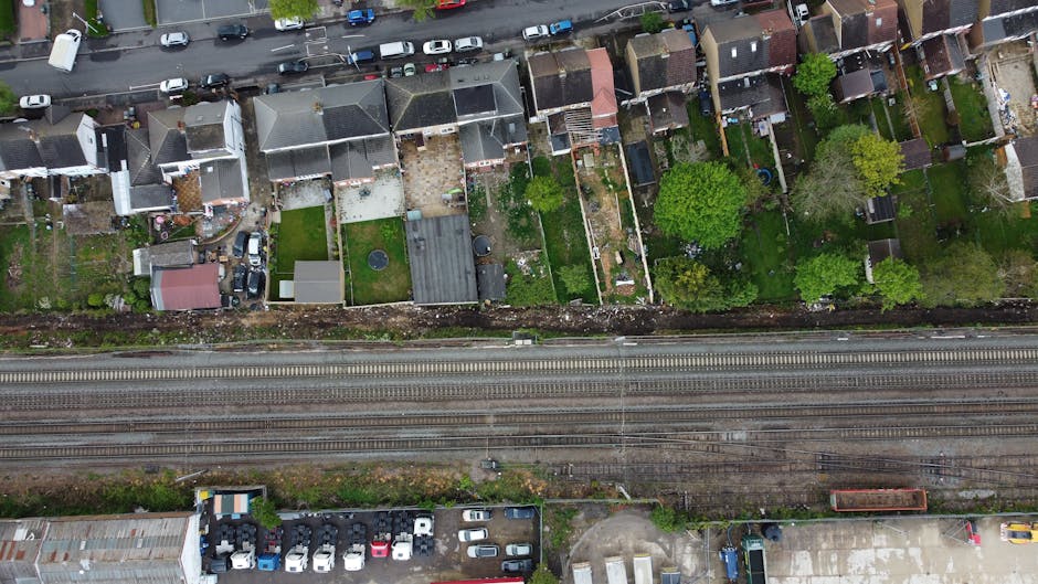 Aerial view of a residential area showing multiple terraced houses with small back gardens, some with patio furniture, sheds, or small lawns. The properties are separated by fences, with trees and greenery visible among the gardens. A busy road runs parallel to the row of houses, with parked cars along its length. Below the road, railway tracks extend across the image, indicating a nearby train line. In the foreground, a parking lot filled with various vehicles is situated adjacent to an industrial or commercial building. The scene depicts an urban environment during daylight hours, with natural light illuminating the houses, road, and railway lines, all highlighting the typical layout encountered during house removals and home relocation activities, especially when coordinated by services like Man with Van Herne Hill.