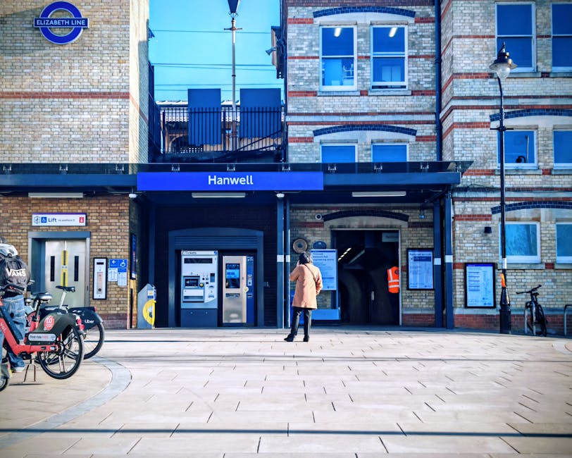 A person dressed in a beige coat and dark pants stands outside the Hanwell station entrance, a brick building with large windows and a blue sign displaying the station name. To the left, there are several bicycles secured to bike racks, including a black bike with a basket and a red bike with stickers, along with a yellow and blue ticket machine. The station entrance features a black canopy with an Elizabeth Line sign positioned above it. On the right side of the entrance, there is a notice board with information and a small poster, and a person in a high-visibility orange jacket is visible inside the station’s ticket area, seen through the open doorway. The surrounding environment includes a sidewalk made of large paving slabs, a black street lamp on the right, and a clear sky above. This scene captures a typical urban setting suitable for home relocation or moving services, as facilitated by companies like Man with Van Herne Hill, while highlighting the station's accessibility for furniture transport and packing and moving logistics.