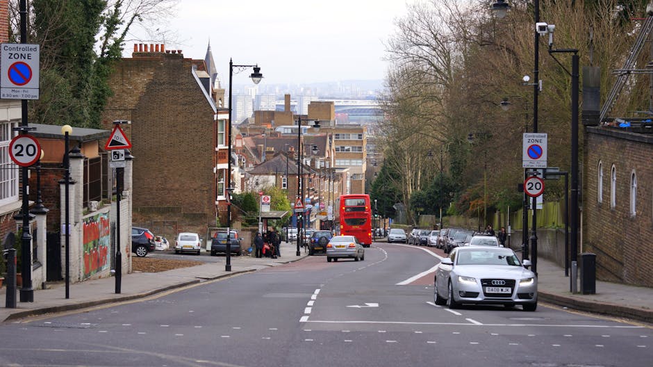 A street scene in Herne Hill during daytime, featuring a curved road with parked cars on both sides and a few moving vehicles, including a prominent silver Audi driving towards the camera and a red double-decker bus traveling away. On the left side, there are brick residential buildings with small front yards and various parked cars, while on the right side, there are additional parked vehicles and a brick wall. Street signs indicating a controlled zone and a 20 mph speed limit are visible, along with lampposts and trees lining the pavement. The background reveals a distant view of an urban skyline with mixed-height buildings and office blocks under an overcast sky. This setting illustrates a typical urban environment suitable for house removals or furniture transport services, with clear space for loading and unloading outside residential properties, aligning with the moving process and logistical considerations for services like those offered by Man with Van Herne Hill.