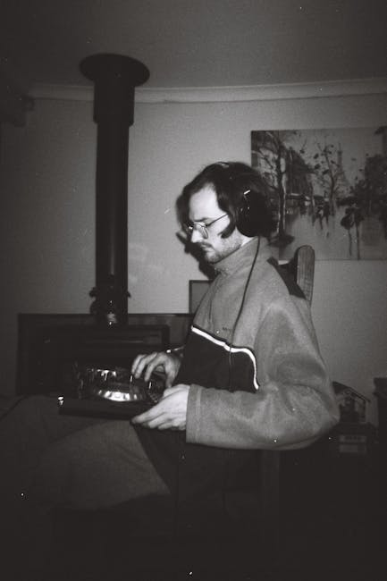 Black-and-white photograph of a man wearing glasses, headphones, and a light-colored jacket, standing indoors near a corner, holding a vinyl record in his hands. He is positioned in front of a wood-burning stove with a visible chimney pipe extending upward. The room features a framed painting of trees hanging on the wall, and various objects including a box and furniture are partially visible in the background. The setting appears to be a living space, possibly during a home relocation or packing process, with the man engaged in handling records amidst other household items. The image captures a quiet moment of packing or organizing, relevant to furniture transport and moving activities carried out by [COMPANY_NAME] for house removals or property relocations.