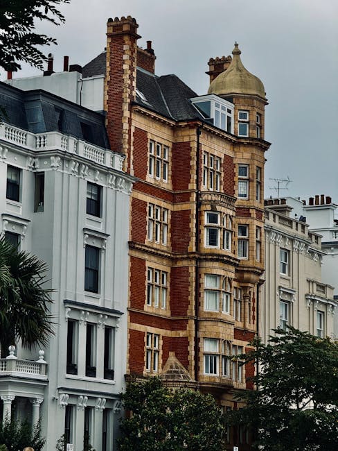 Photograph of a row of historic Victorian-style residential buildings in Herne Hill, featuring ornate brickwork, decorative window frames, and a small cupola on the rooftop. In the foreground, a street scene shows parts of a white building with intricate architectural detailing, and a tree partially obscures the lower section of the buildings. The cloudy sky overhead suggests overcast weather. This urban environment highlights the distinctive architecture typical of Herne Hill, where property renovations often require careful planning and specialist removal services, such as those provided by Man with Van Herne Hill, to facilitate home relocations and furniture transport within these heritage-listed structures.