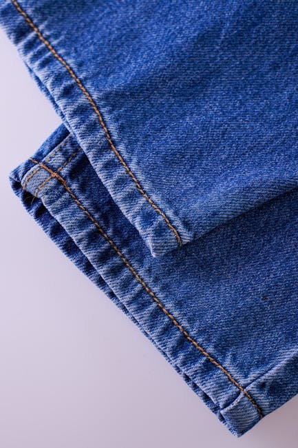 Close-up of a folded pair of blue denim jeans with visible orange stitching, placed on a plain light-colored surface. The material shows typical denim texture with slight creases and faded areas. The jeans are partly inside an open cardboard box filled with white packing paper, suggesting they are being prepared for packing or transportation during a home relocation. In the background, part of a wooden furniture piece or hardwood flooring can be seen, indicating an indoor environment. The overall scene captures the packing process involved in furniture transport and moving services, as provided by Man with Van Herne Hill, focused on street-by-street house removals in Herne Hill, London.