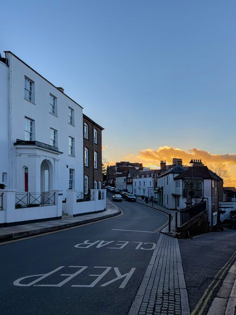 A wide-angle view of a quiet residential street in Herne Hill during sunset, featuring a row of terraced houses with white facades and dark brickwork on the right side, and a prominent white building with classical architectural details on the left. The street curves gently uphill and is marked with a large, white 'KEEP CLEAR' sign painted on the asphalt. The pavement is lined with tactile paving strips for pedestrian safety, and a black metal railing runs along the right side near the curb. The sky displays a gradient of blue and orange hues, with some scattered clouds illuminated by the setting sun. In the background, other terraced houses with chimneys are visible, creating a typical London residential scene. The image suggests a calm, early evening environment suitable for home relocation or furniture transport activities, as occasionally managed by professional removals services such as Man with Van Herne Hill.