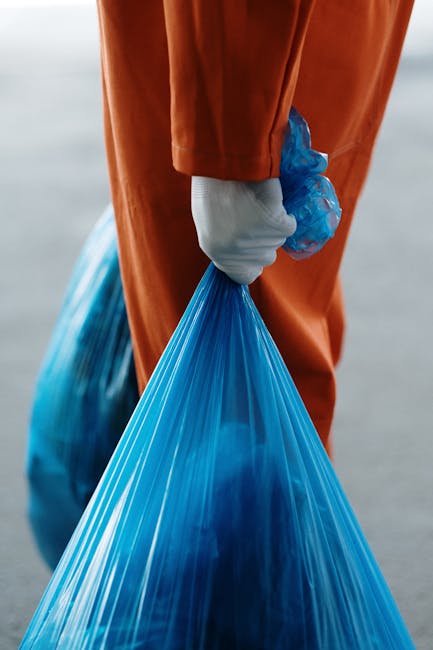 A close-up of a person wearing an orange jumpsuit and white gloves, holding a blue plastic rubbish bag filled with waste. The background is blurred, suggesting an outdoor setting. This image relates to waste disposal during household relocations, which is an important aspect of moving and clearance services. The individual is positioned to the side, with the bag hanging downward, emphasizing the handling of waste materials such as packaging or other refuse, potentially involved in house removals or clearance tasks. The scene illustrates the careful handling of waste as part of the packing and decluttering process prior to a move, relevant to the services offered by Man with Van Herne Hill and the compliance with waste regulations in Lambeth related to home relocation. The lighting is natural, highlighting the details of the gloves, bag, and jumpsuit, aligning with professional moving practices.