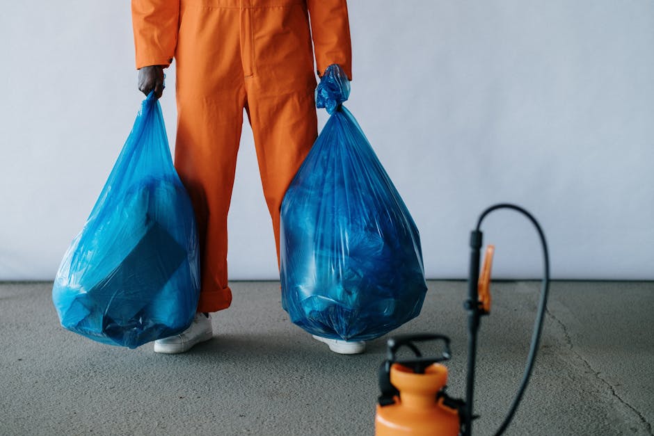 A person wearing orange work overalls and white shoes is standing indoors on a grey carpeted floor, holding two large blue plastic garbage bags filled with waste or packing materials, with the bags hanging from both hands at each side. The background shows a plain light grey wall, and in the foreground, there is an orange and black handheld sprayer or pump. The scene is related to packing, loading, or waste disposal during a home relocation or furniture transport process, illustrating steps involved in removal services offered by Man with Van Herne Hill, as part of their removals service category. The image highlights the disposal of unwanted packing materials during a house move, supported by visible materials and equipment used in removal logistics.