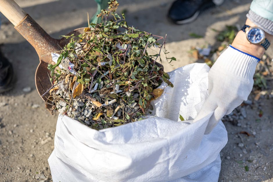 A close-up of a person wearing an orange jumpsuit and white gloves, holding a blue plastic rubbish bag filled with waste. The background is blurred, suggesting an outdoor setting. This image relates to waste disposal during household relocations, which is an important aspect of moving and clearance services. The individual is positioned to the side, with the bag hanging downward, emphasizing the handling of waste materials such as packaging or other refuse, potentially involved in house removals or clearance tasks. The scene illustrates the careful handling of waste as part of the packing and decluttering process prior to a move, relevant to the services offered by Man with Van Herne Hill and the compliance with waste regulations in Lambeth related to home relocation. The lighting is natural, highlighting the details of the gloves, bag, and jumpsuit, aligning with professional moving practices.