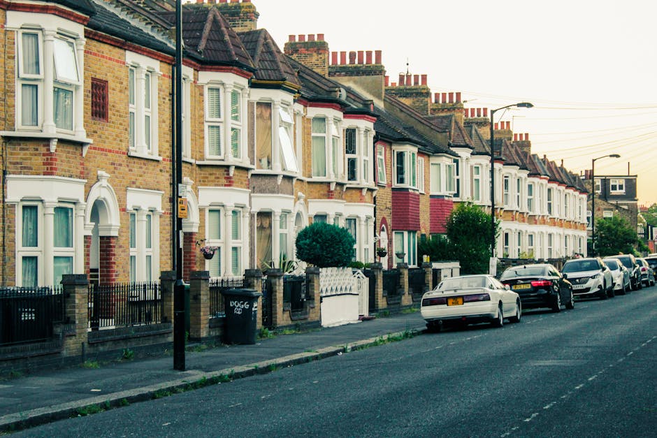 A row of terraced Victorian-style houses with brick façades and white-framed bay windows on a residential street in Herne Hill during daytime. The pavement in front of the houses has small front gardens enclosed by metal fences, and some yards feature potted plants and small bushes. Several cars are parked along the curb, including sedans and hatchbacks. In the foreground, a black moving trolley is visible on the sidewalk next to a large cardboard box and a plastic wrapping material, indicating preparations for a home relocation. The scene appears well-lit with natural daylight, and overhead power lines run parallel to the row of houses, emphasizing the typical London suburban setting. This image relates to house removals and moving logistics, as it captures the environment where professional removal services like those provided by Man with Van Herne Hill operate, ensuring efficient furniture transport and packing during property relocations.